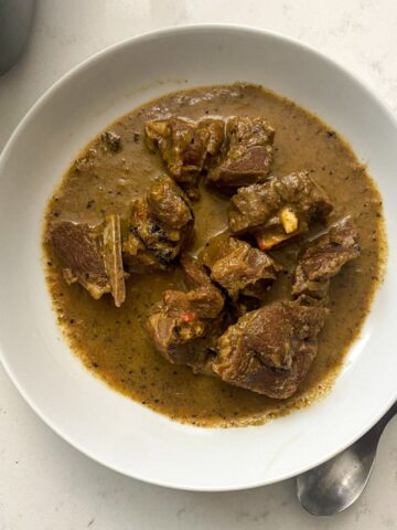 this is a picture of goat meat pepper soup in a white bowl on a marble counter top with a silver spoon